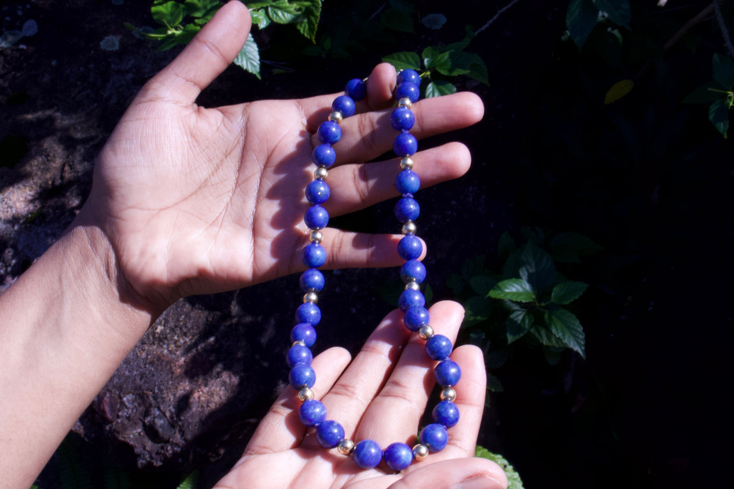 Hands holding a blue beaded necklace against a dark, natural background