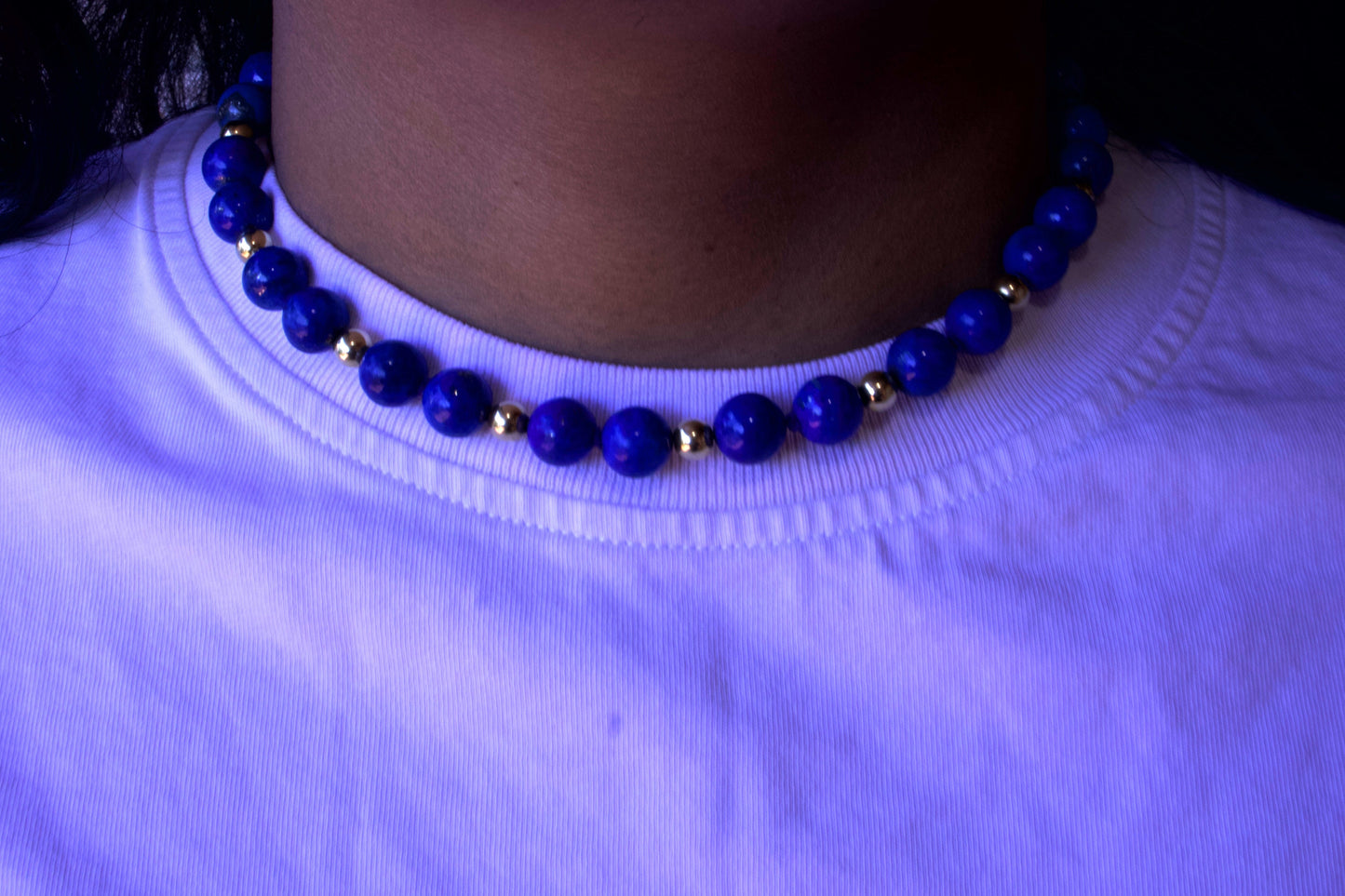 Person wearing a blue beaded necklace on a white shirt with a dark background