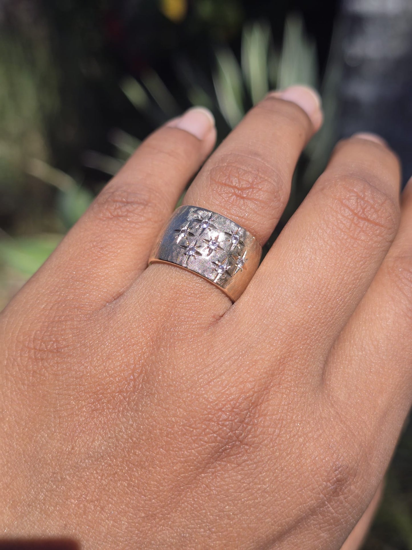 Hand wearing a gold ring with a textured band against a blurred natural background