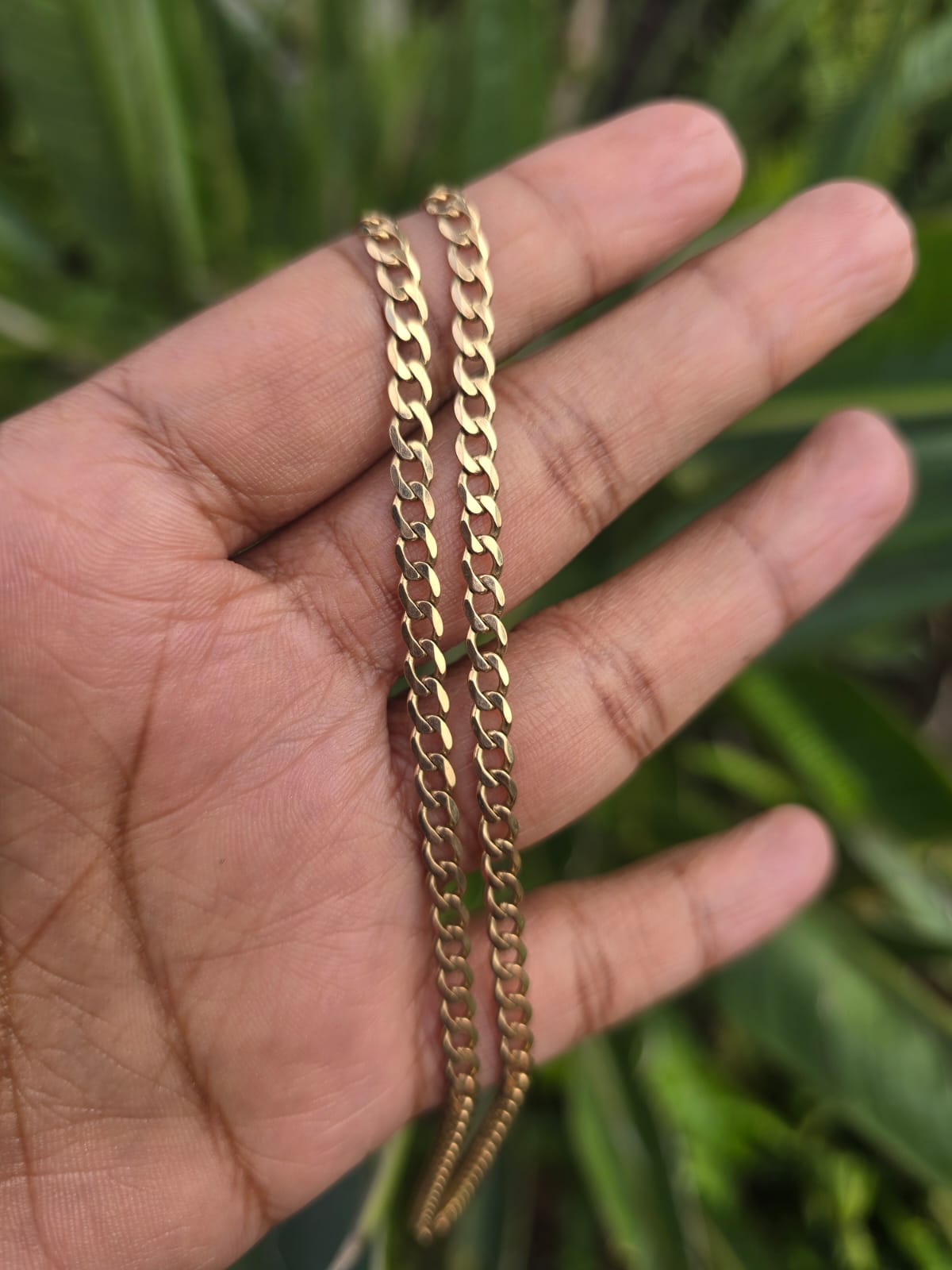 A gold-toned curb chain necklace draped over a person's hand, with a blurred green plant background.