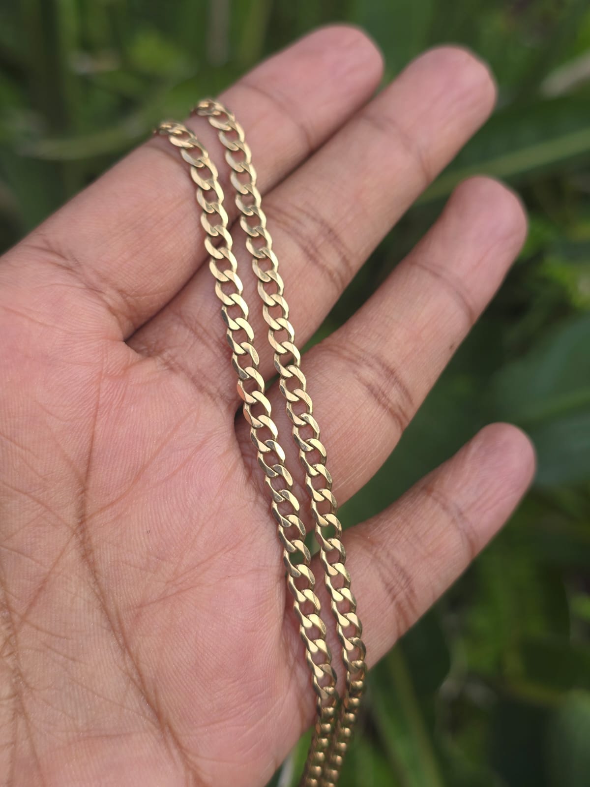 A close-up of a hand holding a gold-toned curb chain necklace against a blurred green outdoor background.